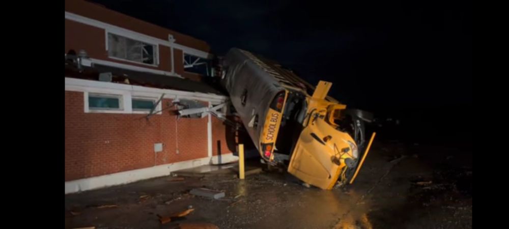 A photo of a school bus picked up and smashed onto the lower roof of a brick High School.
