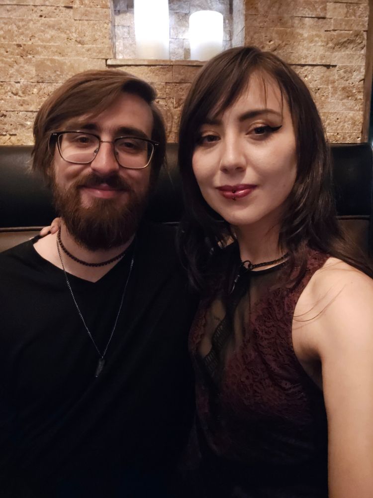 a couple in a booth - man on right is wearing a black long sleeve with a choker and long necklace, has glasses, short brown hair, and a beard. I am on the right with long dark hair and a (partially visible) lace/mesh dress !!!