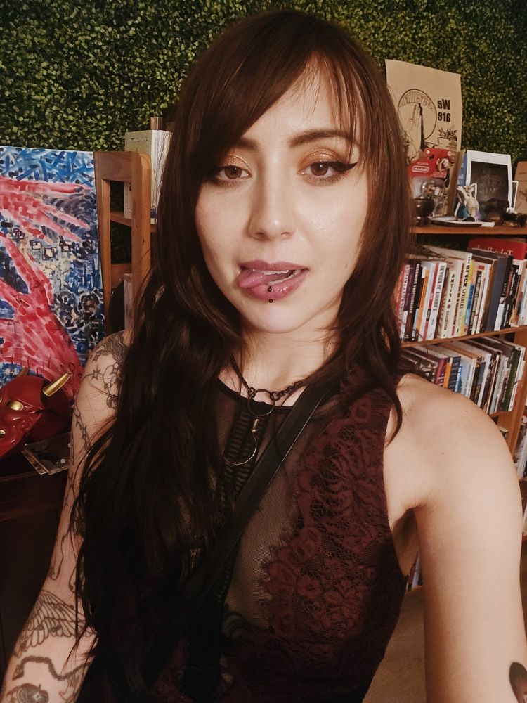 an androgynous human with long dark hair and makeup on in front of books and a leafy wall ++ art. the human is sticking their tongue out ! they are wearing a lace/mesh dress with a black scythe necklace. 
