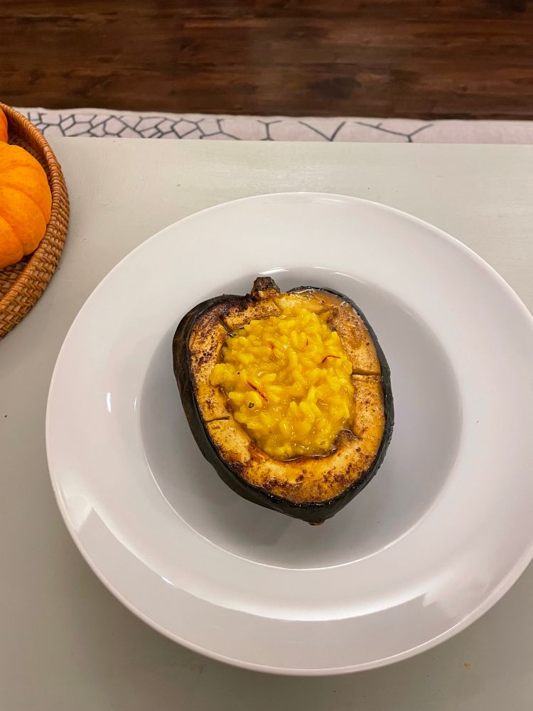White dish holding an acorn shaped squash, black skin on the outside, orange in the middle. Inside is risotto with saffron strands, almost the same colour as the squash.