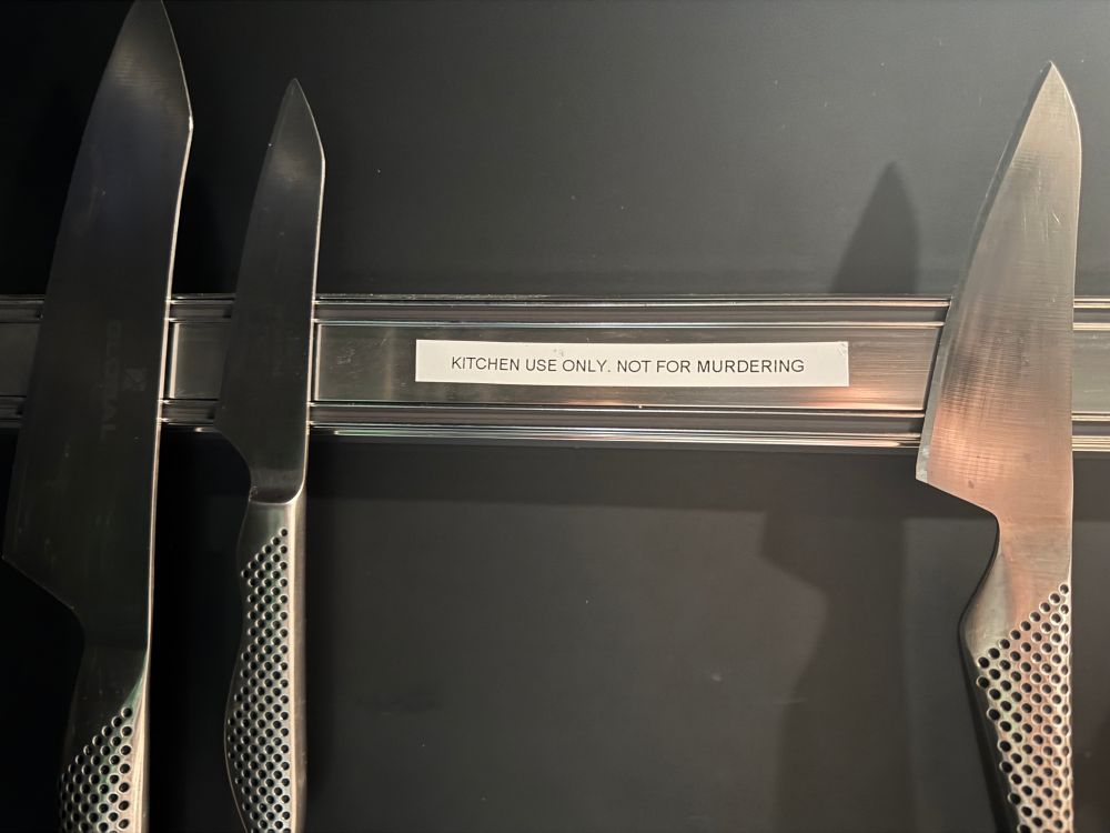 A photo of a kitchen magnetic knife board with some knifes and a label that says: “kitchen use only, not for murdering”