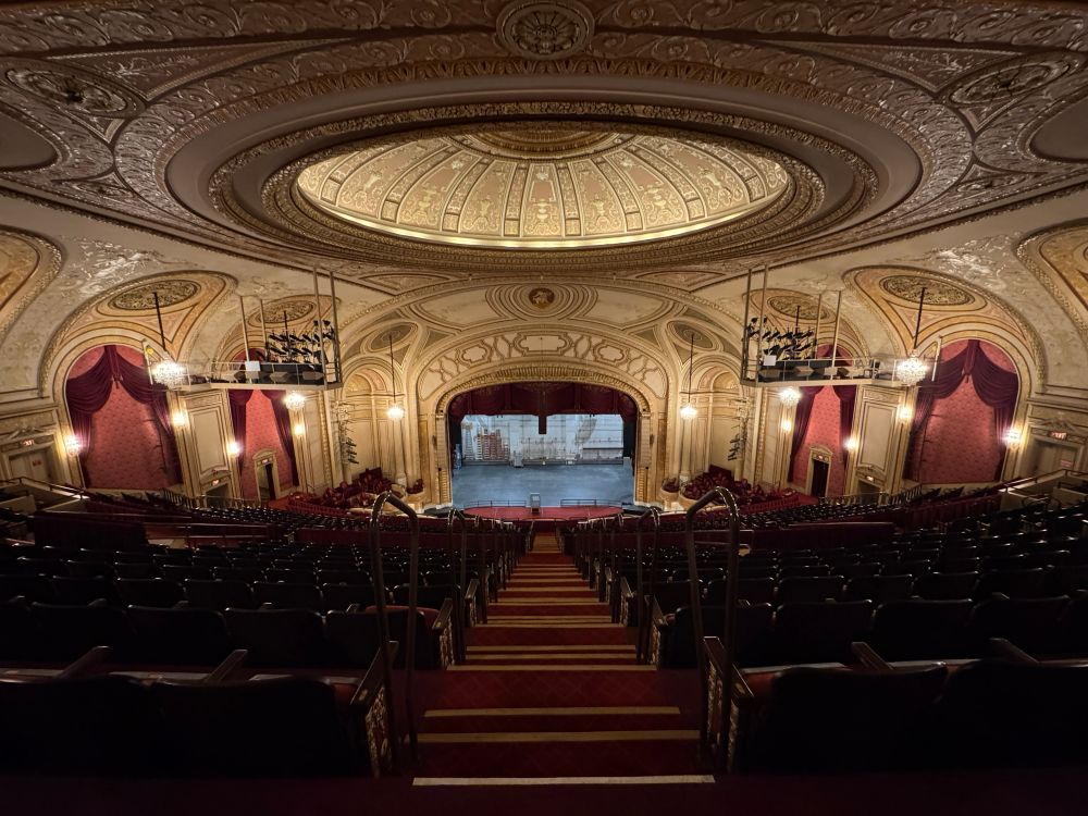 A photograph of the Palace Theatre at Playhouse Square in Cleveland, OH. 
The picture centers on an empty stage. In the foreground is the audience seating for the theatre. 
Red curtains hang over the box seats of the theatre ornamented with intricate patterns from the theatre’s Vaudeville beginnings. 
The room is illuminated by chandeliers and light boxes on the ceiling, creating a golden and warm atmosphere.