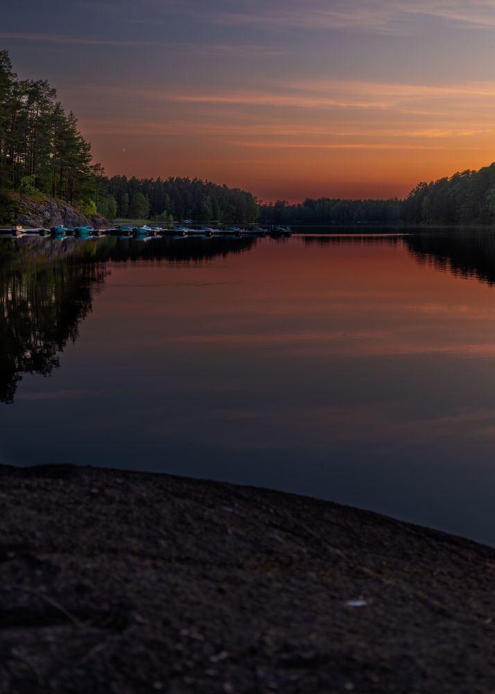 A serene lakeside scene at sunset, with the sky painted in hues of orange and purple. The calm water reflects the vibrant colors of the sky and the surrounding trees. A row of small boats is docked along the shore, and the silhouette of a forest is visible in the background. The foreground features a rocky shoreline, adding texture to the tranquil landscape.