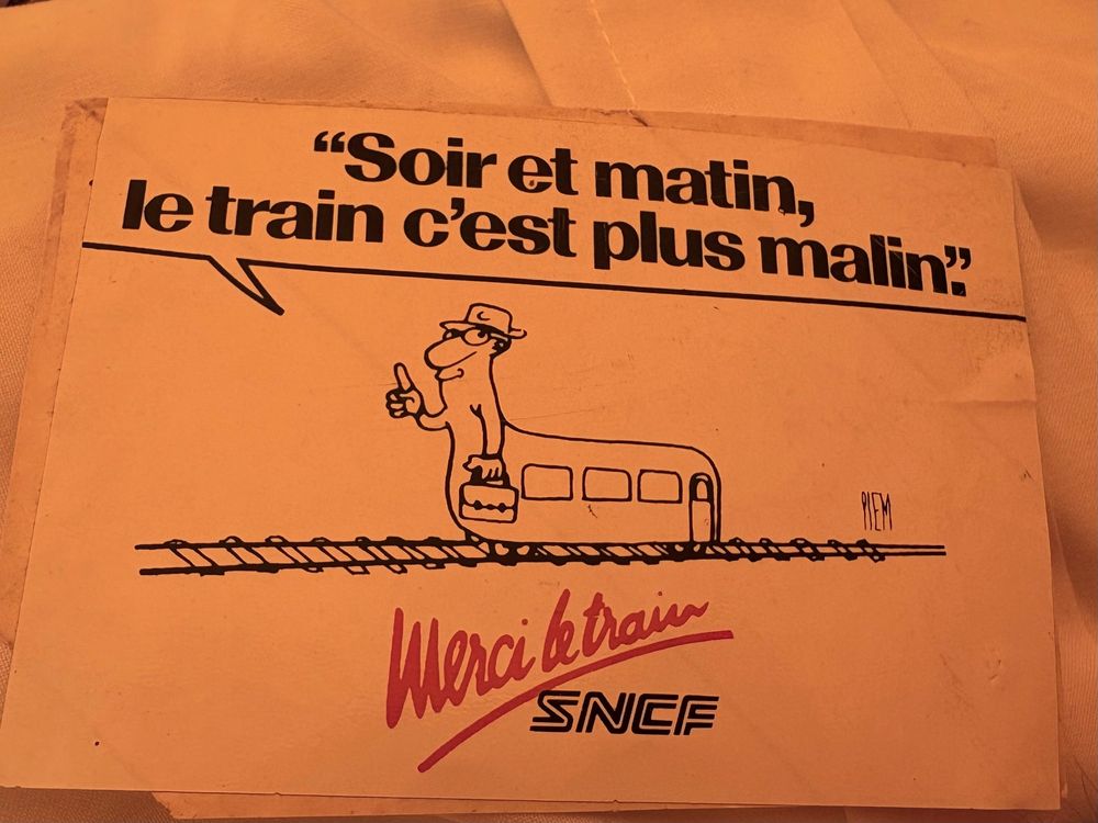 Autocollant posé sur un drap. Le fond est blanc, un train personnifié portant un chapeau et une sacoche, roule sur des rails. Le train dit « soir et matin, le train c’est plus malin ». Avec un slogan en rouge « merci le train » juste au dessus du logo SNCf.  