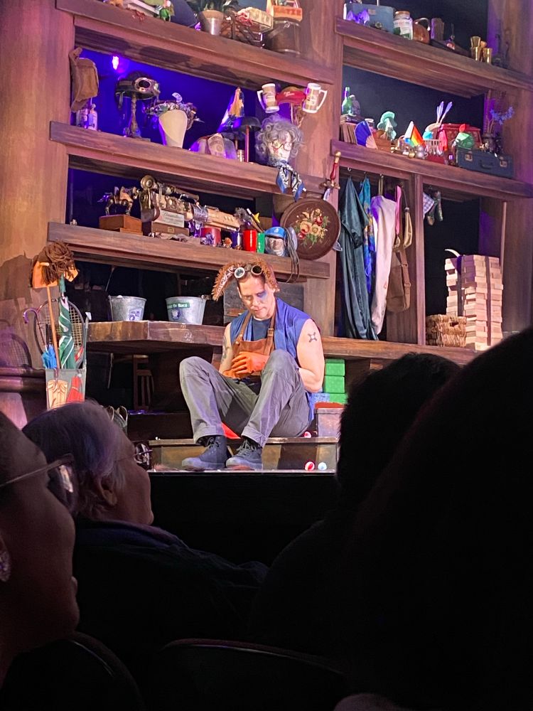 Actor Neil Newbon, wearing rabbit ears and orange gloves, stares solemnly into the floor of a stage. 