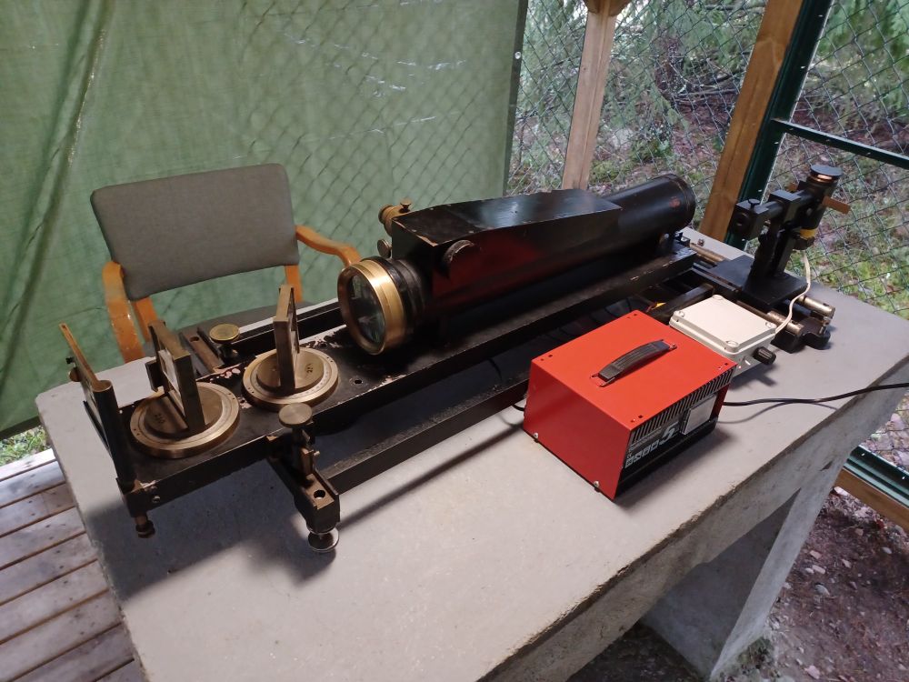 Väisälä comparator box with white light source on table. Mirrors on the left to adjust the light beams. Chair for observer behind the table. Under roof surrounded by metal fence.