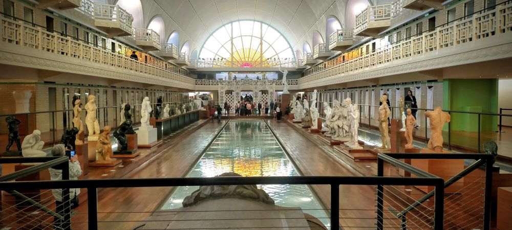 Salle principale du musée de la Piscine de Roubaix. On peut voir un bassin entouré de statut et les balcon du 1er étage qui font le tour de la salle. Au fond, le célèbre vitrail en demi-cercle représentant le soleil avec un style art-déco. 
