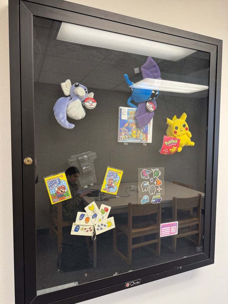 A glass display case with vintage Pokémon plush toys (including Pikachu and Dratini), Super Mario Bros. 3 cards, Among Us stickers, Pac-Man-themed tags, and other gaming memorabilia. A person is reflected working at a table behind the case.