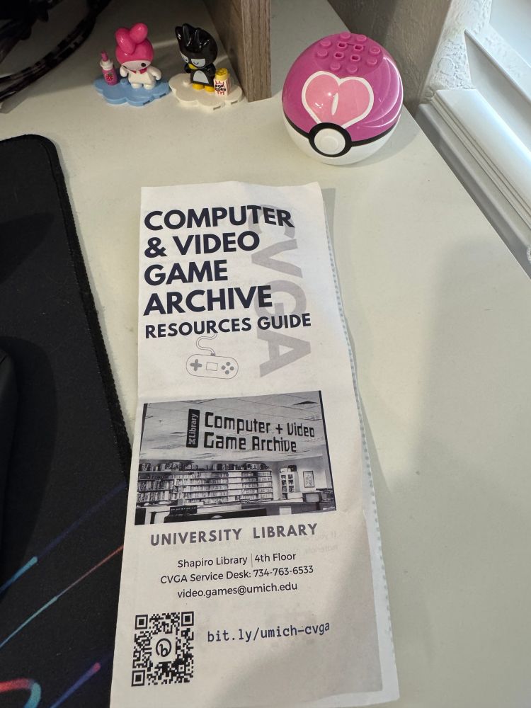A printed flyer for the University of Michigan's Computer & Video Game Archive (CVGA), resting on a desk. The flyer includes a photo of the archive, a QR code, and contact info. In the background are figurines of Hello Kitty characters and a pink Poké Ball speaker.
