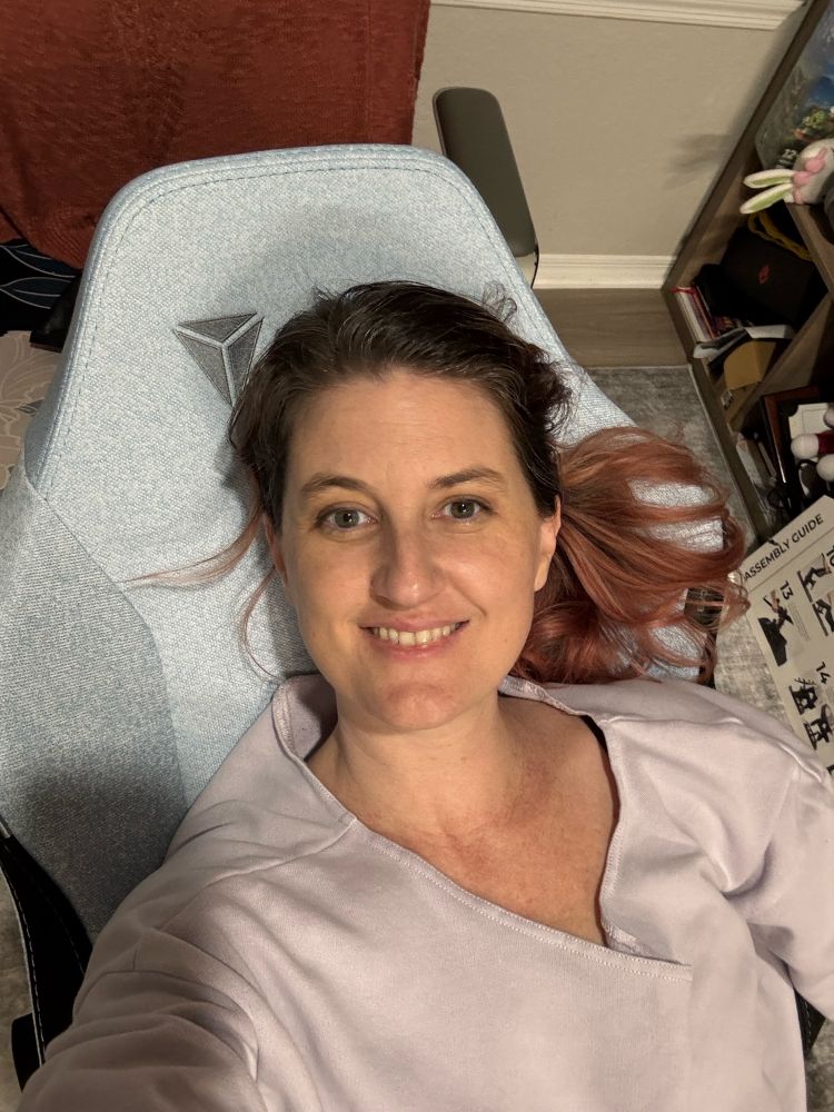 A woman reclining comfortably on a light gray Secretlab gaming chair, smiling at the camera. She is wearing a soft lavender top, and her pink-tinted hair flows slightly to the side. Behind her, a rust-colored sweater is draped over a nearby chair, and a bookshelf filled with colorful items and books is partially visible. The relaxed setting highlights a cozy and welcoming atmosphere.
