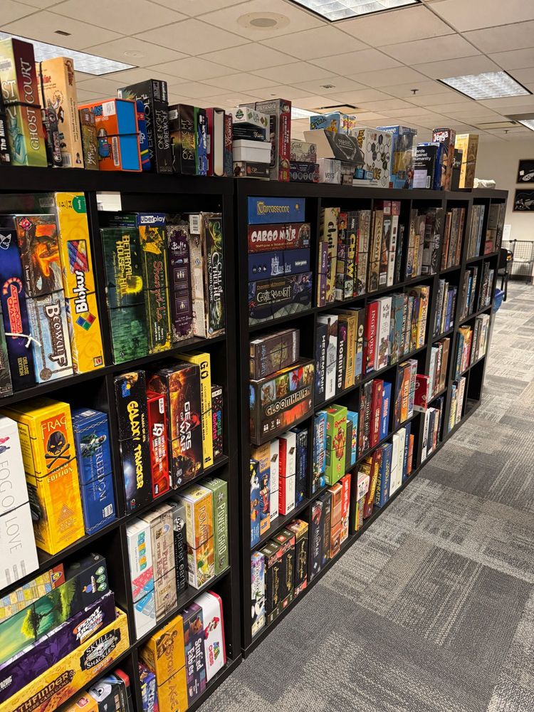A well-organized library of board games on black bookshelves in a brightly lit room. Game titles include Carcassonne, Power Grid, Galaxy Trucker, Pandemic, and more. The floor is carpeted, and the space resembles a community or library game room.