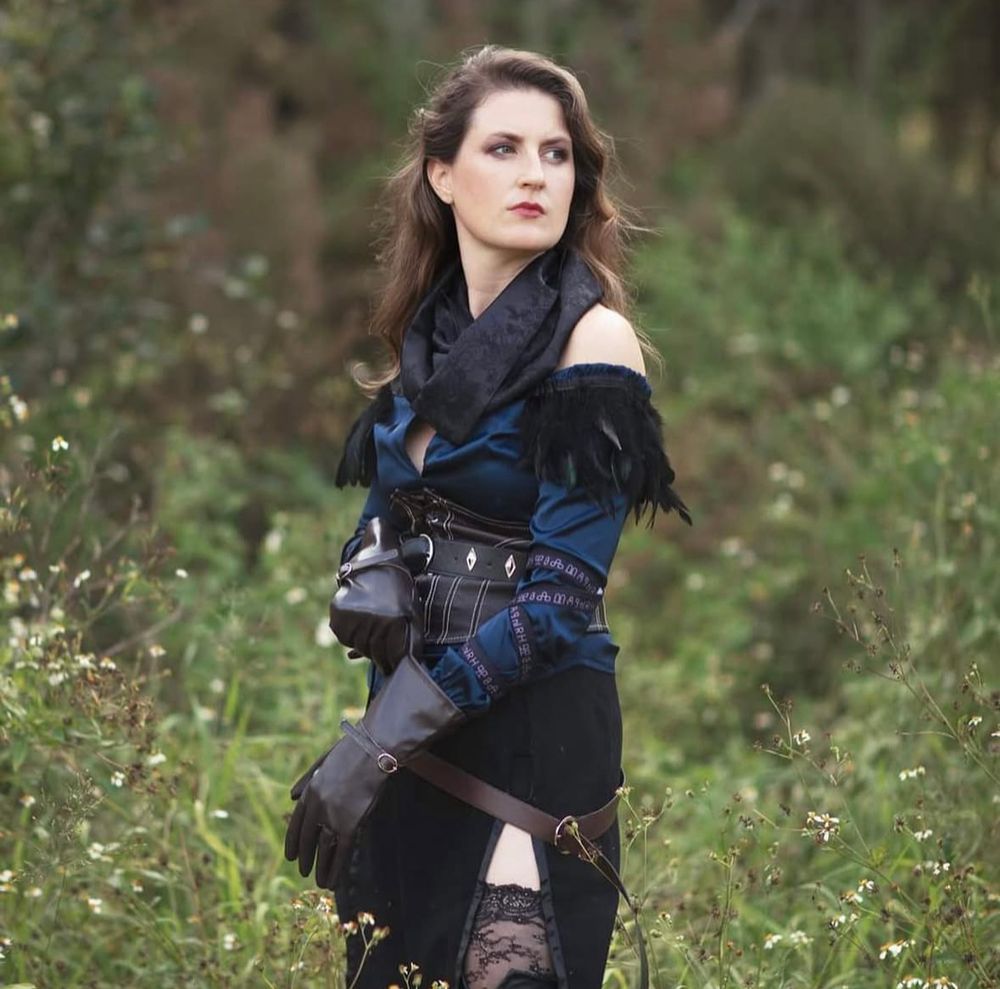 woman with fair skin and long, wavy brown hair stands in a lush, green outdoor setting with small white flowers. She wears a fantasy-inspired outfit consisting of a dark blue off-the-shoulder blouse with black feathered trim, a black corset with silver accents, long black gloves, and a black skirt with a thigh-high slit revealing lace stockings. She gazes into the distance with a confident expression.