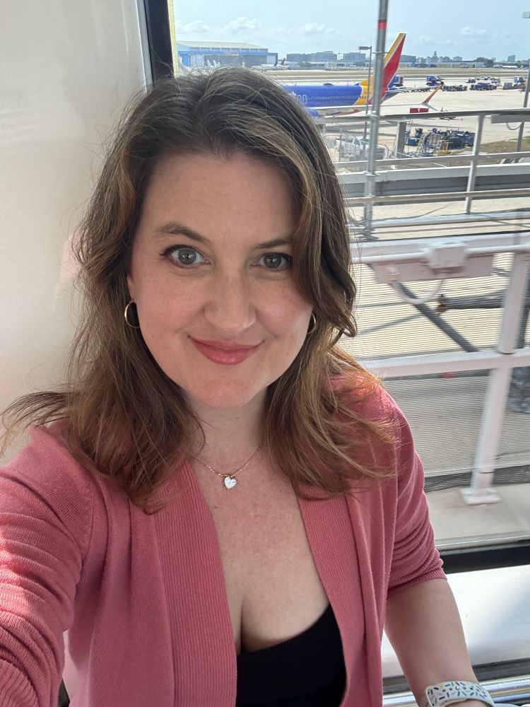 Dr. Kelly McNeil smiling in a selfie at the airport, wearing a pink cardigan with planes and a Southwest Airlines aircraft visible through the window in the background