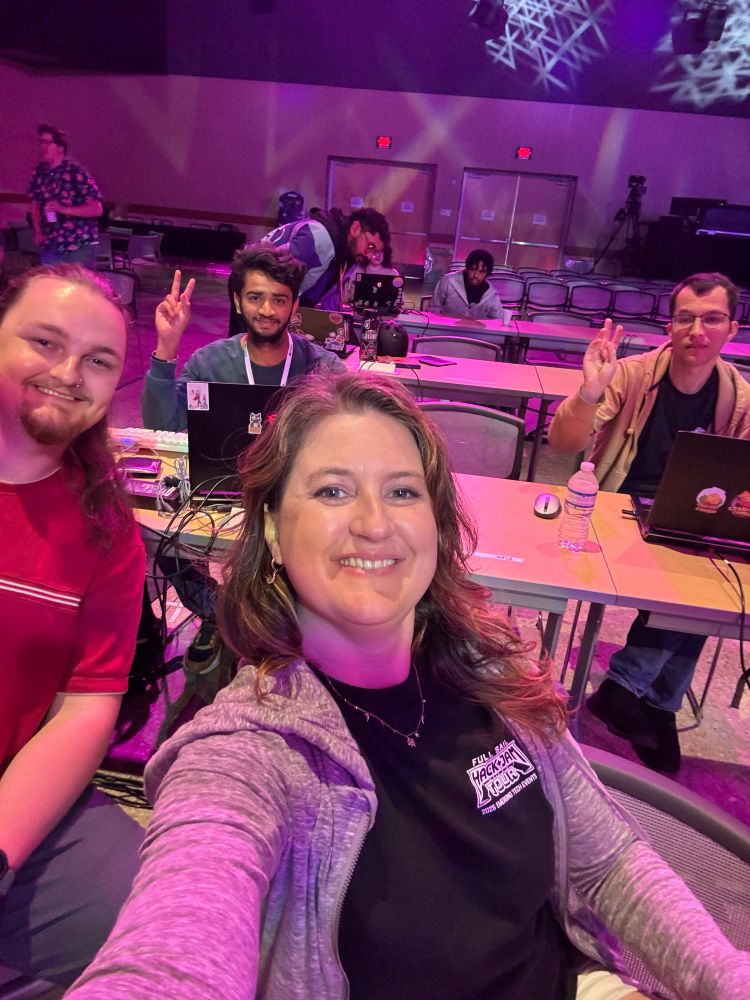 A selfie taken at the Unreal Fest Game Jam with a woman in the foreground smiling and three teammates seated behind her at a row of tables, all working on laptops. Two teammates flash peace signs, and the room is lit with stage lights and spotlights, giving a festive, energetic atmosphere. Other developers are seen working in the background.