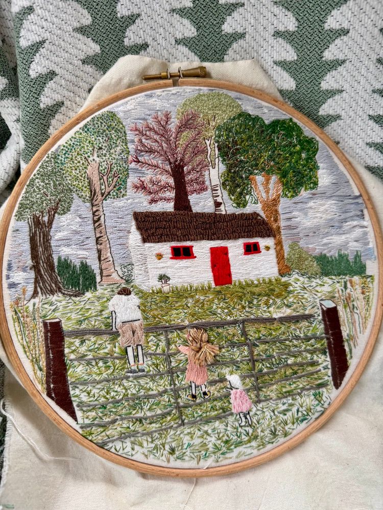 embroidery hoop, hand-stitched with an Irish cottage scene (white washed walls, bright red door & red window trim, surrounded by embroidered trees and three young children climbing a fence to get over to the cottage). 