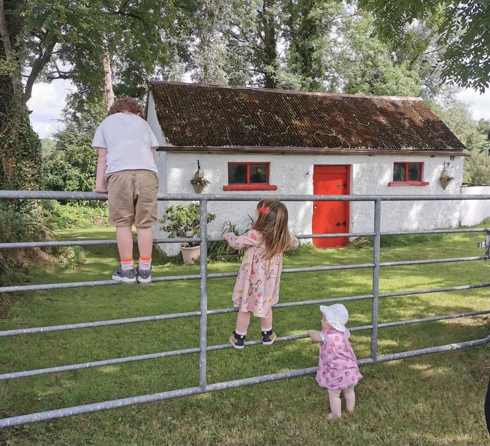 Photograph that embroider is based on. Three young children climb a metal fence in the foreground. Beyond the fence is a whitewashed cottage with red door and windows, surrounded by trees. 