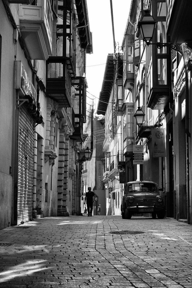 Eine Straße in Palma, Mallorca. Ein Mann läuft eine Straße entlang. Ein Kleinwagen parkt im Schutz der Häuser. Grelles Licht dringt in die Gasse, der gepflasterte Boden liegt im Schatten. Das Bild ist schwarzweiß, kontrastreich, harte Konturen. 