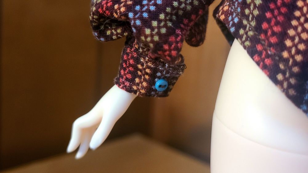A close up photo of the cuff of a doll shirt. The shirt is made of colourful striped fabric. The puffed sleeve is gathered into the cuff, which has a small blue button as a closure.