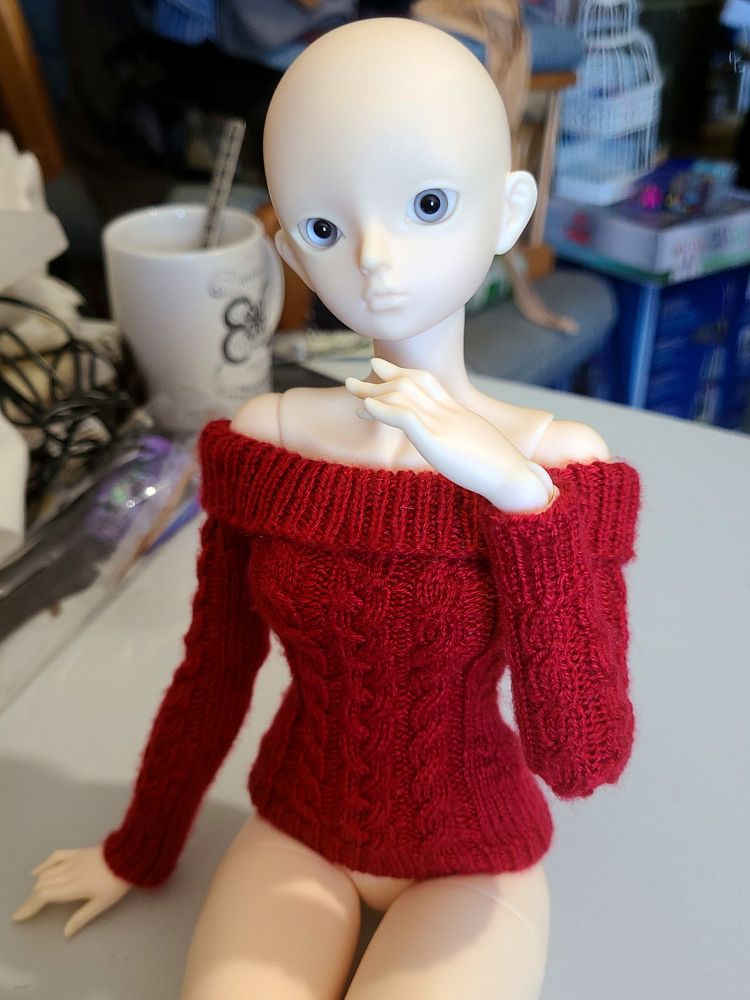 A ball-jointed doll wearing a knitted red off-the-shoulder sweater with cable details.