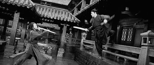 Long haired man with a spear duels a man in a hat in the courtyard of a Chinese temple 