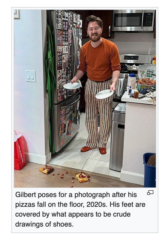 Gilbert poses for a photograph after his pizzas fall on the floor, 2020s. His feet are covered by what appears to be crude drawings of shoes.