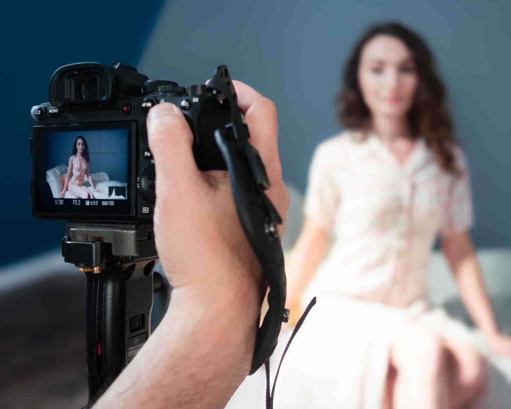 A out-of-focus model poses on a white chaise longue in a studio setting, wearing a plaid dress. In the foreground, in focus, a hand grips a camera where we see the same model in the same pose, but without any clothing.