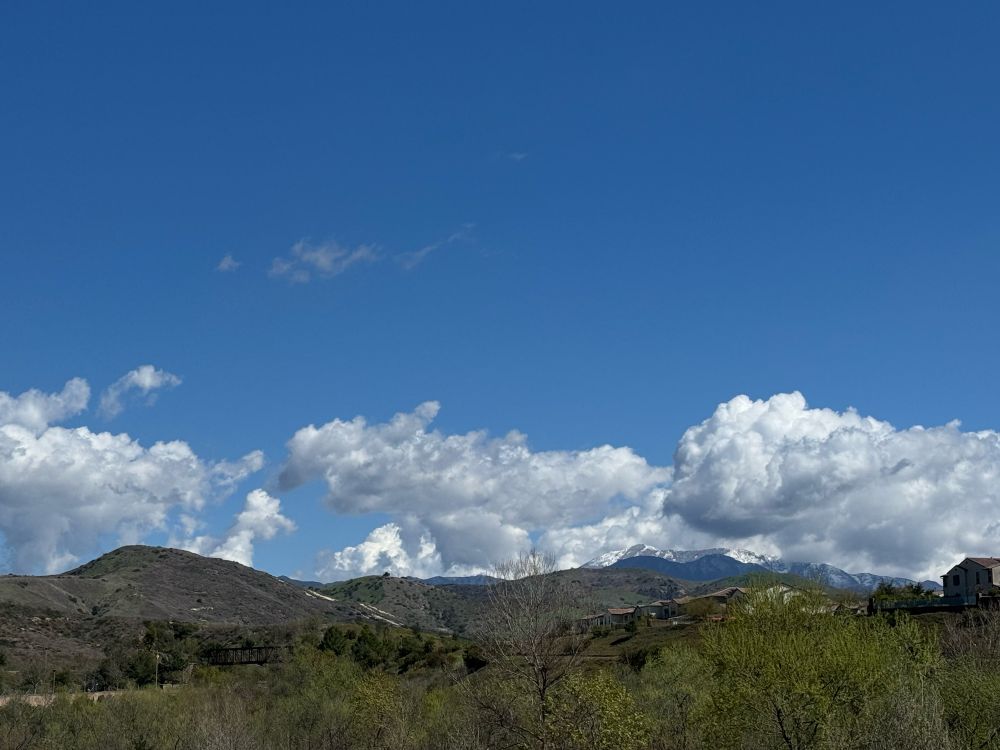 Green brush in the foreground with green and brown hills in the background. There is a blue sky with white clouds hovering over the hills. In the distance behind the hills is a white snow-capped mountain. 
