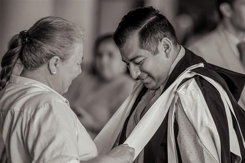 This image shows a black and white photograph of two individuals engaged in a personal moment. One person, appears to be receiving assistance from another person, who is adjusting or tying their graduation gown. 