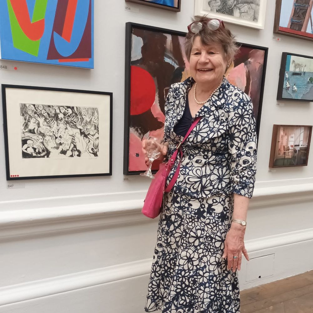 A woman wearing a black and white dress with a flower pattern stands near a wall in a gallery. She holds a glass of water and carries a pink handbag. On the wall are several painting and prints.