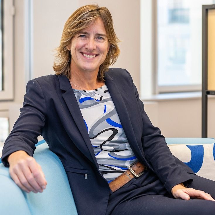 A young woman wearing a blue jacket and a top with curly blue, black and grey lines on it, sits on a sofa. She looks relaxed and smiles at the camera.