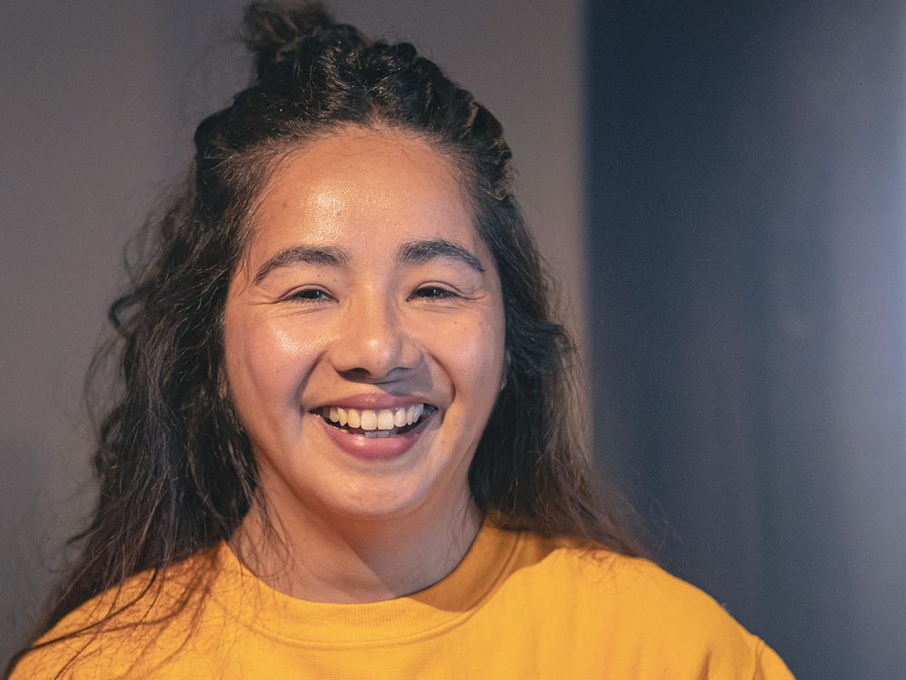 Araceli smiling, wearing a yellow sweatshirt, standing against a grey background 