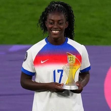 A young woman wearing an England football shirt stands on a pitch and she holds a trophy in her hands.