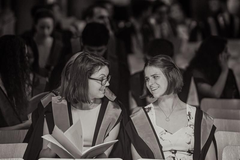 Black and white picture of two graduates, dressed in graduation down, smiling and engaging in a conversation.