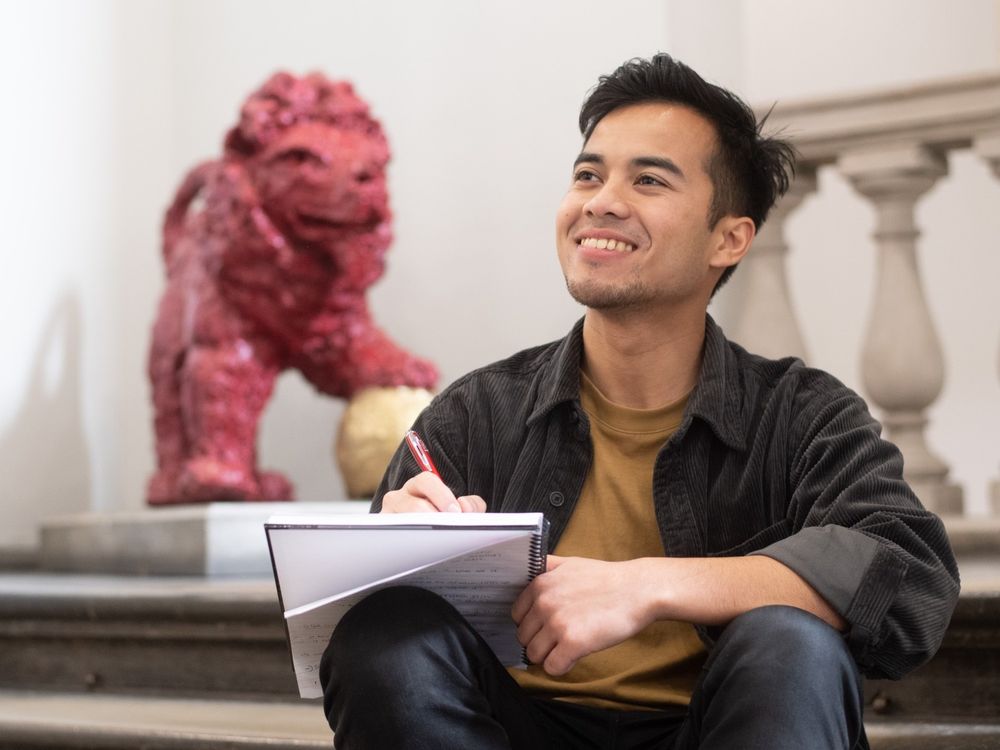 A young man sits on the main stairs inside the King's Building on The Strand. He is casually dressed and writing on a notepad. In the background is the red statue of the King's College London mascot, Reggie.
