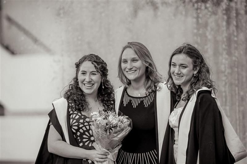 A black and white picture of three graduates in graduation gowns, smiling warmly for a picture,