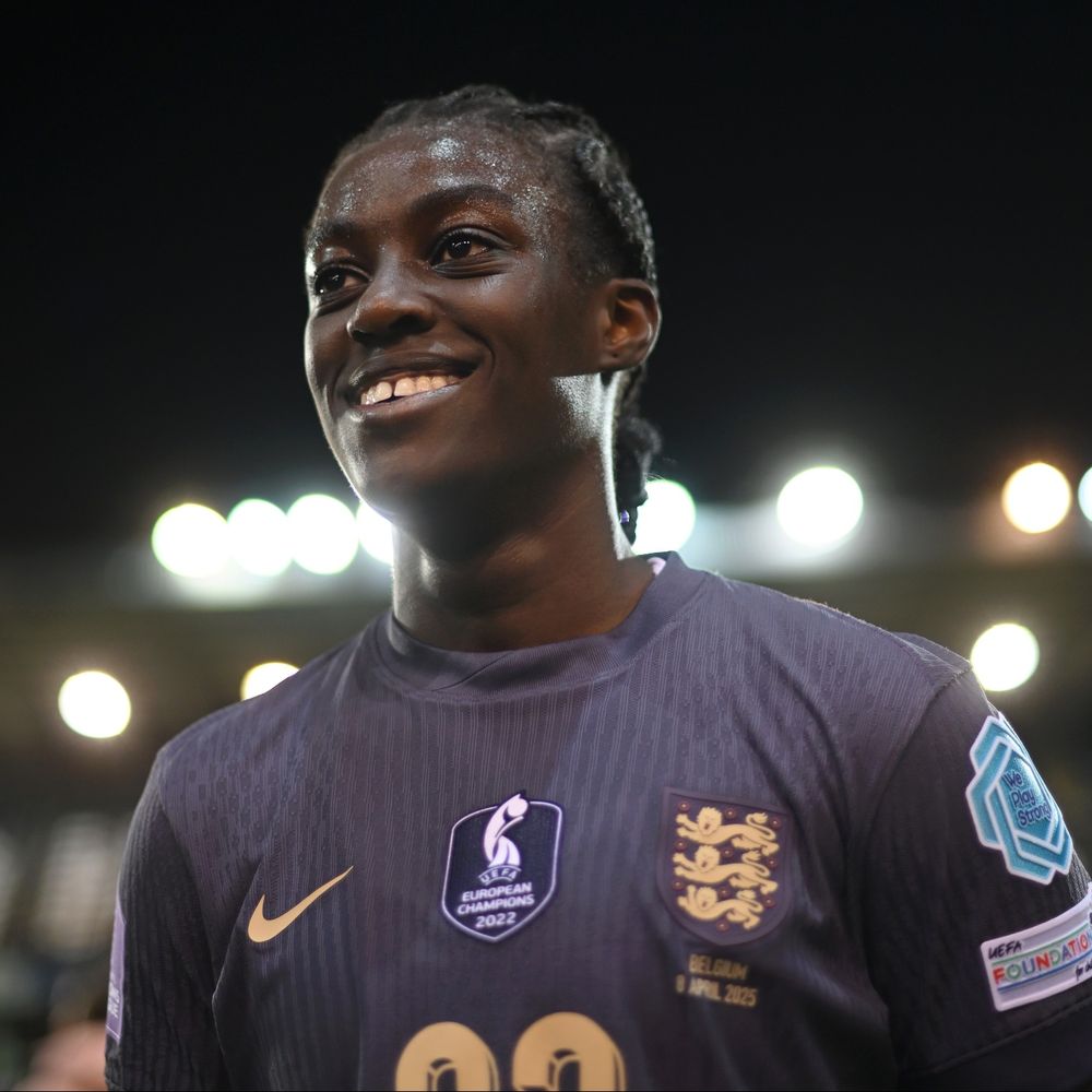 A young woman wearing an England Football Team shirt stands smiling on a football pitch.