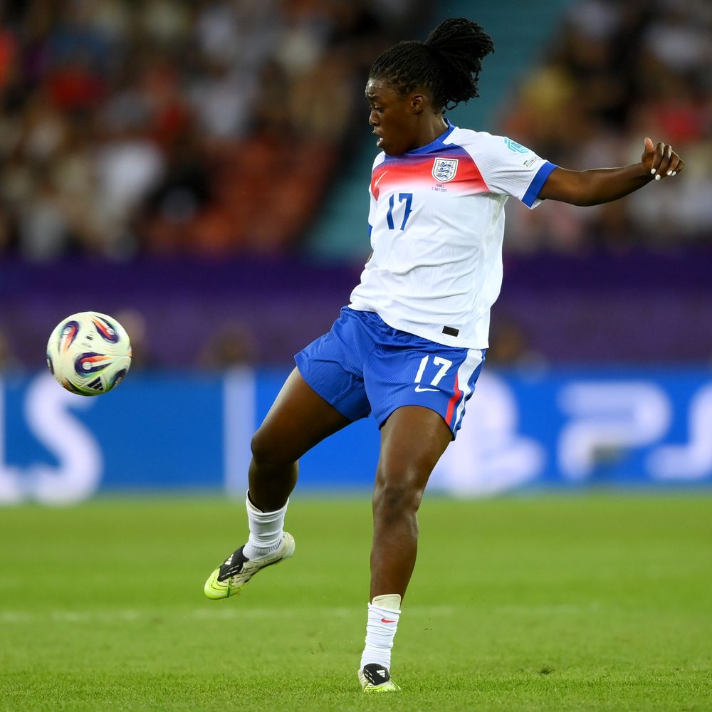 An action shot of a young woman wearing an England football shirt standing on a pitch with the ball coming towards her feet.