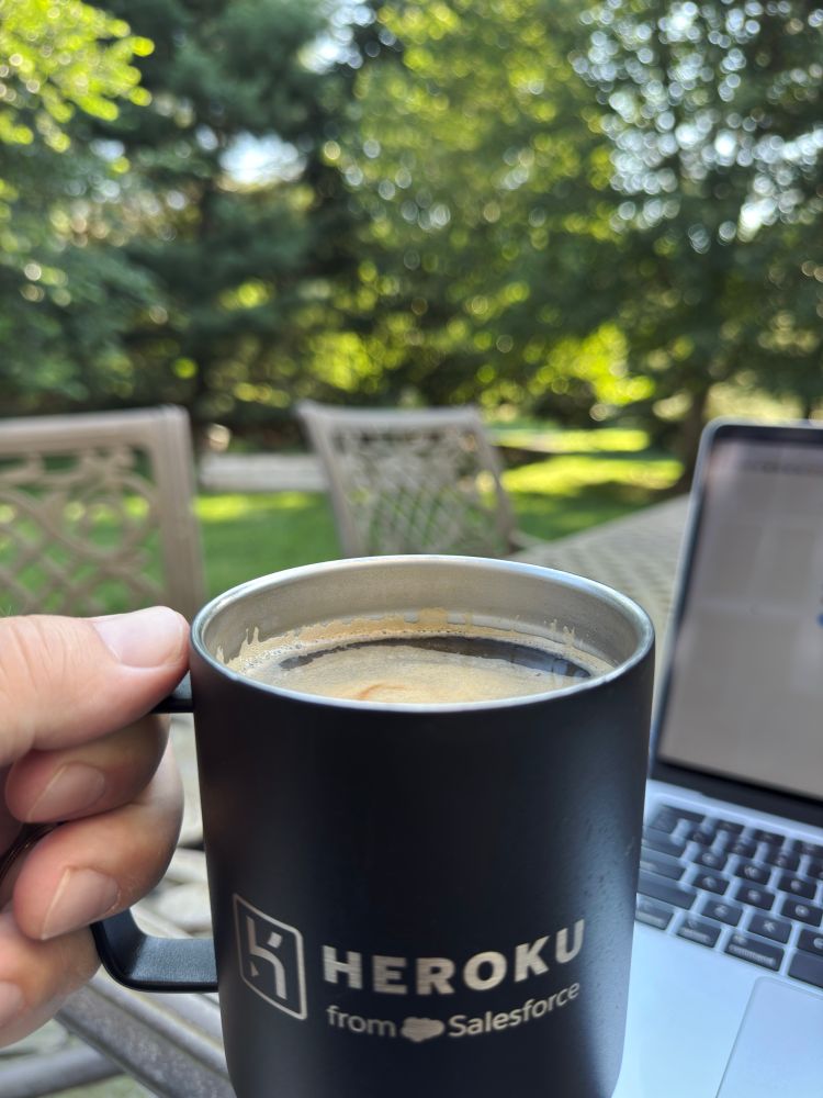 A person holds a black coffee cup in their left hand outside. A computer is open along the right side. Green trees are in the background.