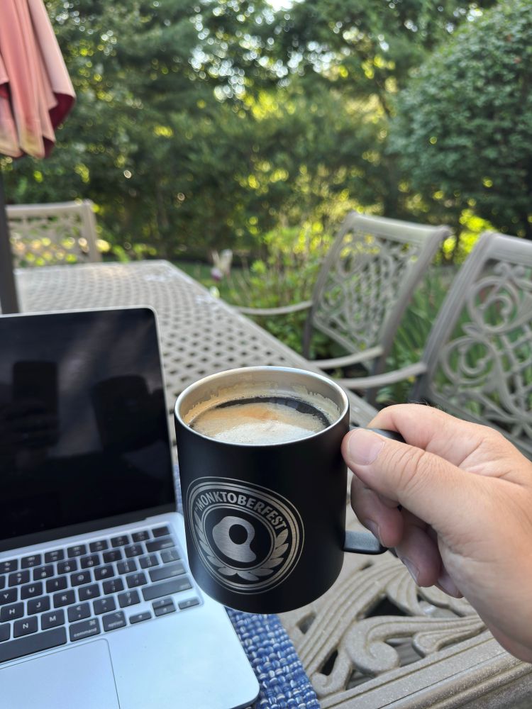 A person holds a black coffee cup in their right hand. A computer sits in the left corner on a blue mat. The person is outside and you can see green trees in the background.