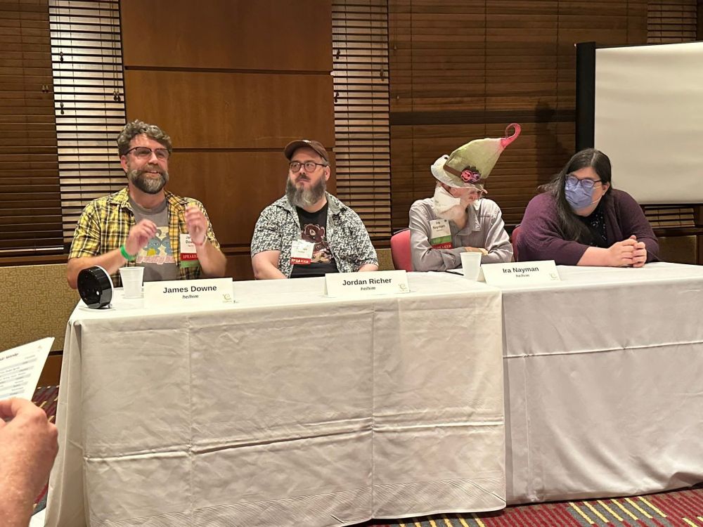 4 people having a disscussion, sitting behind a table. 1 person, not me, is wearing an incredible hat.