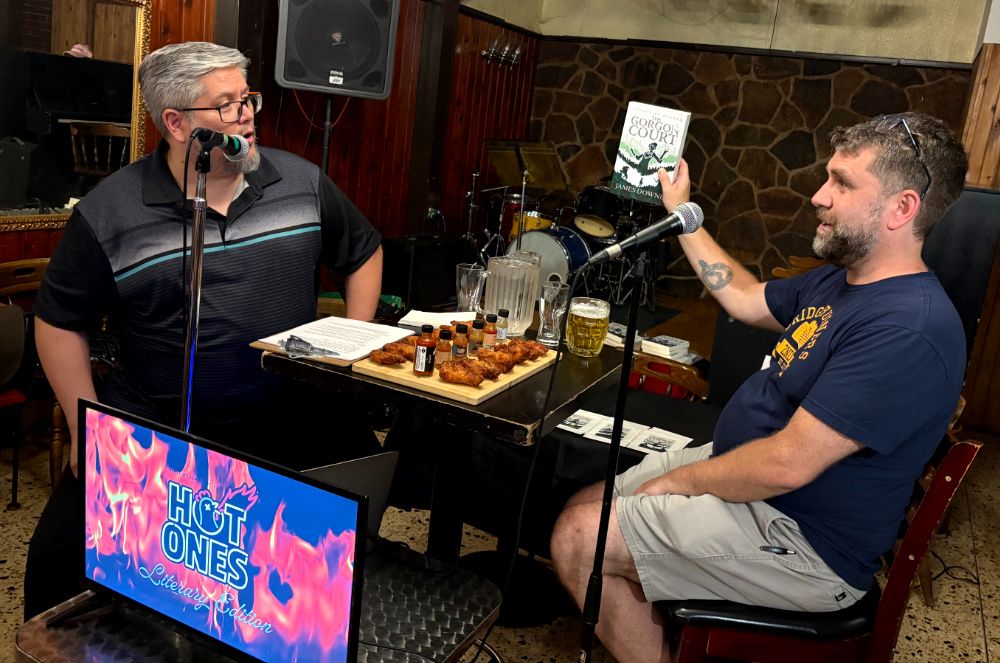 Two men (me and my friend Peter) sitting at a table with hot wings and microphones. The one on the right (me) is holding up a book called THE GORGON COURT.