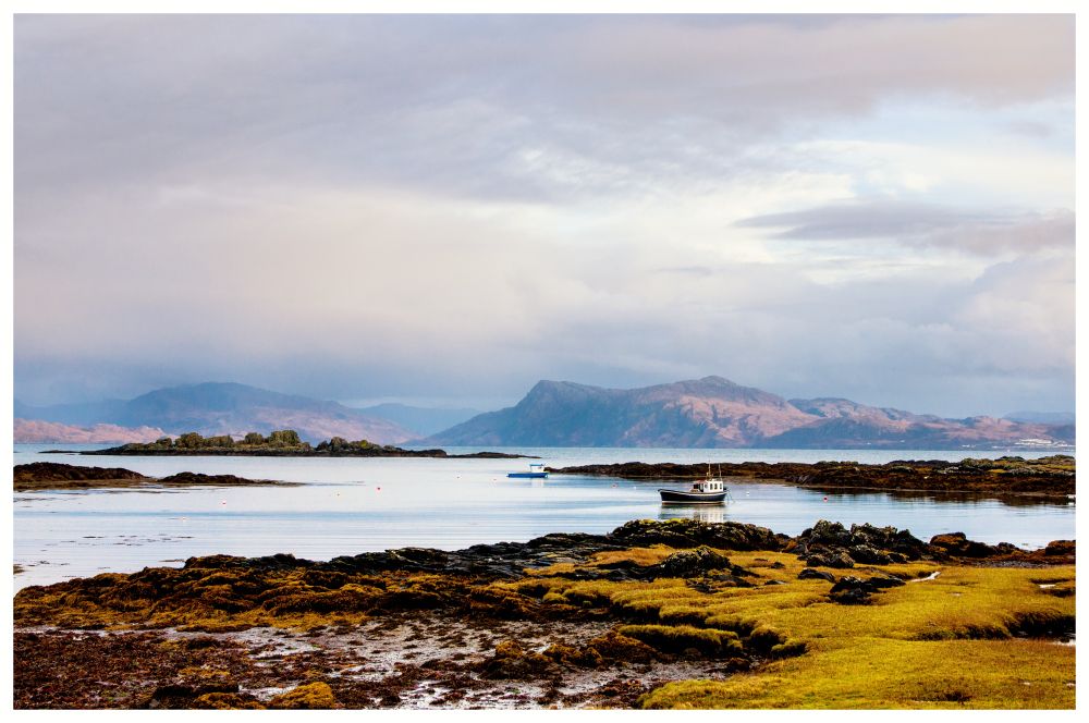 There is a small fishing boat at anchor I. The natural harbour of inlet waterways between the islands and the mainland. There are hills the other side and a cloudy overcast sky. 