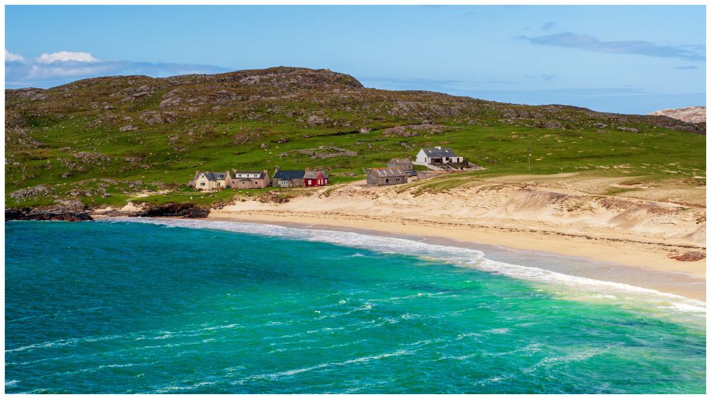 Turquoise sea takes up the lower half of the picture as its rolls in left to right onto a golden sandy beach. At the end of the beach in the middle of the picture is a few small houses and a grassy headland and above a clear blue sky. 