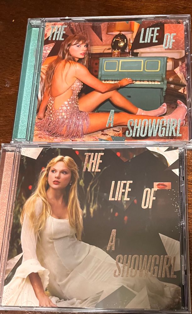 Taylor Swifts “The Life of A Showgirl” album CDS. The top CD features Taylor sitting next to a blue tiny piano wearing a sheer pink, bedazzled dress. The bottom cd features Taylor sitting, looking to the side. She is wearing a white dress with greenery behind her. Both feature the words “The Life of a showgirl” over the picture. 