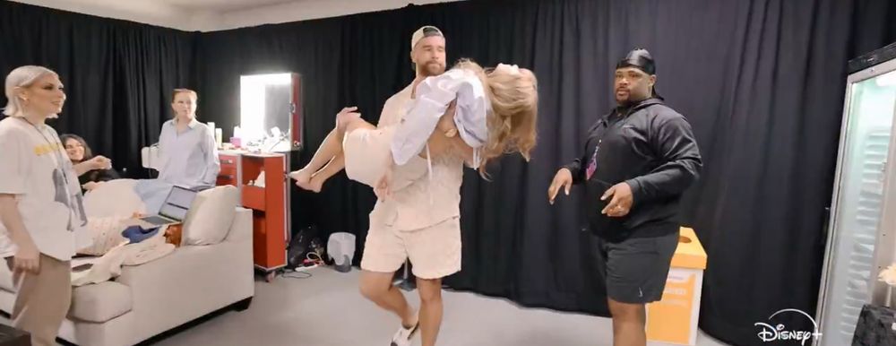 Travis Kelce, in a matching tan shirt/short set, holding Taylor swift, who’s wearing casual clothes. They are in a dressing room of sorts with other people and black curtains around them.