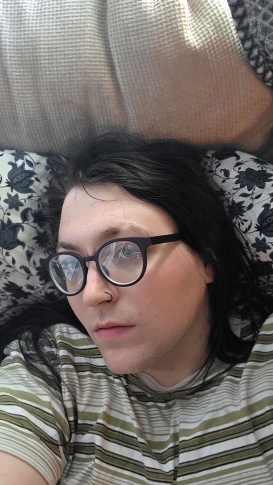 Gretch lays on a bed in a striped shirt her think black glasses and black hair 

