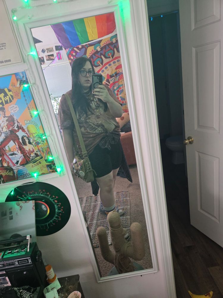 Gretchun stands in the mirror, bike shorts, canvas purse, flannel around her waist, eagle shirt, hair down, shes back on back leg a bit
