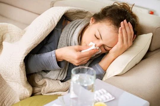 Lassie lying down covered in a blanket holding a tissue to her nose, hand on her head. Pills and a glass of water next to her.