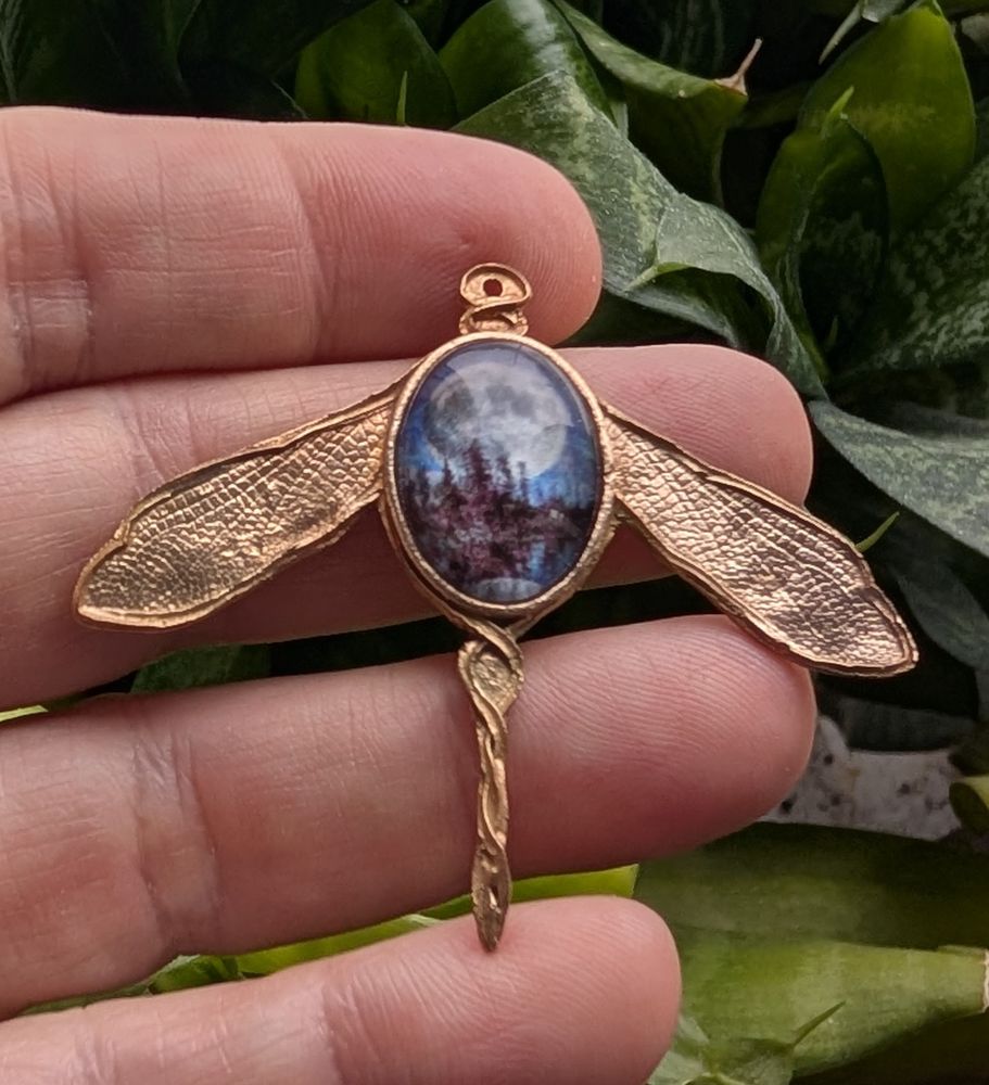 a front view of a bronze pendant in the shape of a damselfly, with a glass cabochon featuring an image with trees and the moon.