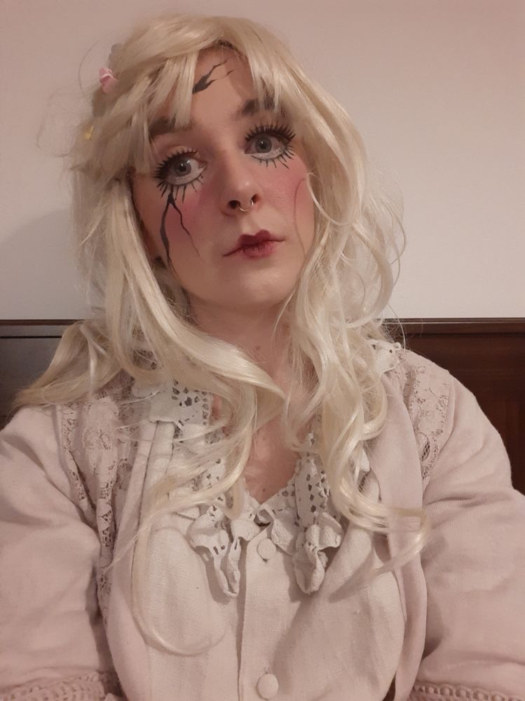 Selfie von Merri. Sie trägt eine blonde Perücke, eine helle Bluse mit Spitze, eine rosa Strickjacke und hat Make up im "broken doll" Stil aufgetragen (große Augen, angeklebte Wimpern, rote Lippen und schwarzer Kajal als Bruchstellen). Sie starrt abwesend in die Kamera.